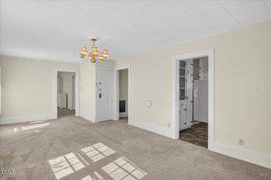 A spacious, carpeted living room with cream walls, white trim, and a ceiling light fixture. Two doorways lead to other rooms, one showing part of a white kitchen with tile flooring. Sunlight streams in through unseen windows.