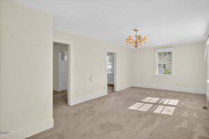 Unfurnished room with beige carpet, off-white walls, two doorways, a window letting in natural light, and a brass chandelier centered on a white ceiling.
