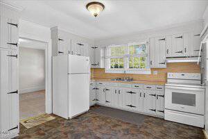 A kitchen with white cabinets and black hardware, a white refrigerator, a white oven with a range hood, a double sink under two windows, wood backsplash, and tile flooring. An adjacent room is visible through a doorway.
