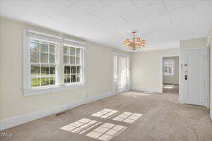 A bright, empty room with beige carpet, cream-colored walls, large double windows, a white door, a small chandelier on the ceiling, and sunlight casting window shadows on the floor.