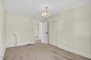 A small, empty room with beige carpet, cream-colored walls, a white ceiling, and a brass chandelier. There are two doors, a wall outlet, and a washer or dryer hookup visible on one wall.