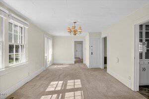 A carpeted room with cream-colored walls, large windows letting in natural light, a ceiling light fixture with multiple shades, and several doorways leading to adjacent rooms.