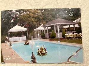 A backyard pool with two floating floral arrangements, a gazebo, white folding chairs, and tents set up in the background, suggesting an event setup. Trees and greenery surround the area.