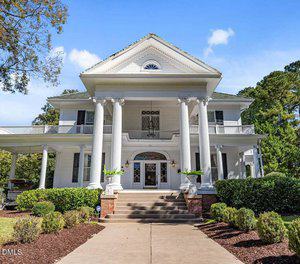 Elegant large white, two-story house at 221 Gilliam Street with prominent columns, a wraparound porch, symmetrical windows, decorative trim, and a brick staircase. Well-kept shrubs line the walkway of this Oxford NC 27565 gem.