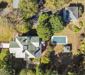 Aerial view of a large house with a green roof, a driveway, trees, landscaped yard, a covered rectangular pool, garden beds, and a detached building nearby.