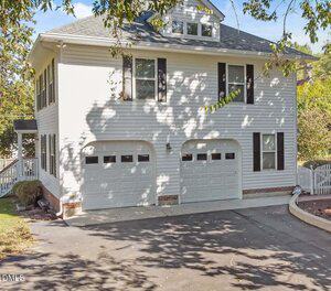 Two-car garage attached to a white house with black shutters, gray roof, and a paved driveway. Trees cast shadows on the driveway and exterior, and there is a small fenced yard on the right.