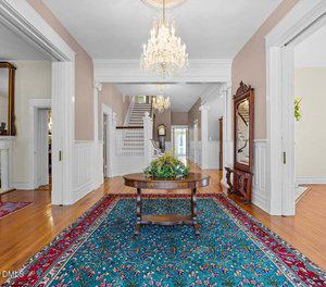A spacious hallway with hardwood floors, a large blue patterned rug, a wooden table with greenery, white paneled walls, a grand staircase, chandeliers, and an ornate mirror. Several doors lead to adjoining rooms.