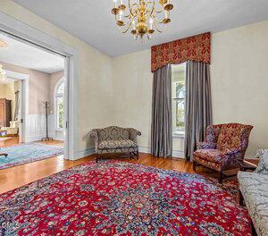 A formal living room with a red patterned rug, two upholstered armchairs, a loveseat, hardwood floors, a brass chandelier, and a window with floor-length curtains and a valance. Another room is visible through an open doorway.