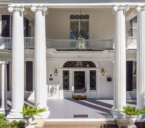 The front entrance of a white, two-story house features four large columns, a balcony with a railing, glass double doors, black lantern-style lights, and potted ferns on either side of the steps.