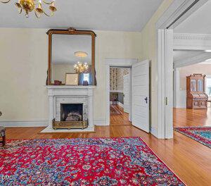 A traditional living room with a white fireplace and large mirror above it, wooden floors, a red patterned rug, a chandelier, and open doorways leading to adjacent rooms with similar decor.