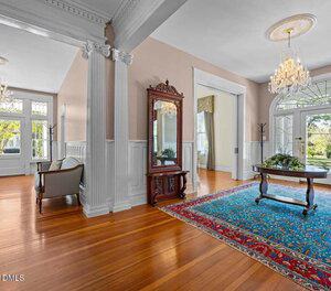A spacious entryway with polished hardwood floors, white columns, a large mirror, a crystal chandelier, a blue and red patterned rug, and a wooden table topped with greenery. Tall windows and double doors let in natural light.