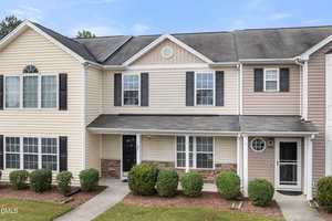 A two-story townhouse with beige and tan siding, black shutters, and a covered entryway. There are trimmed bushes in front of the building and a concrete walkway leading to the doors.