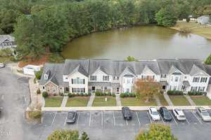 A row of connected townhouses faces a small, calm lake surrounded by trees. Several cars are parked in front of the buildings, and the area appears residential and well-maintained.