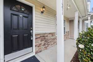 Close-up view of a house entrance with a black front door, lantern-style light fixture, stone and beige siding, a white post, windows, and bushes with white flowers beside the walkway.