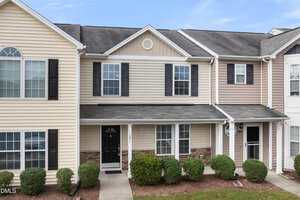 A two-story townhouse with beige siding, black shutters, and a black front door, featuring a covered entryway, trimmed bushes, and a concrete walkway leading to the entrance.