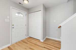 A clean entryway with light wood flooring, a white front door with a window, a white double closet door, light gray walls, and a staircase with a wooden handrail.