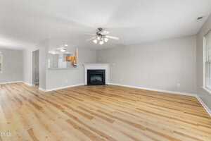 Spacious empty living room with light wood floors, neutral walls, a ceiling fan with lights, a fireplace, and large windows. The kitchen and dining area are visible through an open doorway.