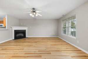 A spacious living room with light wood flooring, a white ceiling fan with lights, a fireplace with a white mantel, light gray walls, and a large window letting in natural light.