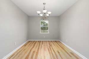 Empty room with light wood flooring, light gray walls, a central window showing greenery outside, and a ceiling-mounted chandelier with five lights. There are white baseboards and electrical outlets on the walls.
