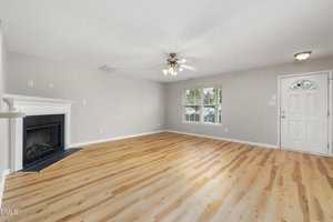 A spacious, empty living room with light wood flooring, a white ceiling fan, a fireplace with a white mantel on the left, a large window, and a white front door on the right. Walls are light gray.