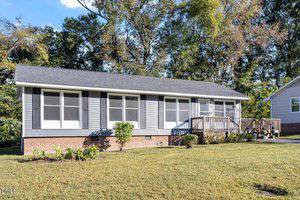 Single-story house with gray siding, white trim, and a brick foundation. The home has several large front windows, a wooden ramp leading to the entrance, and a neatly maintained front yard with shrubs and grass.