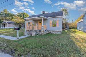 A small single-story house with a front porch, stone steps, and orange shutters, set on a grassy lot with a sidewalk, mailbox, and neighboring homes visible. The sky is partly cloudy.