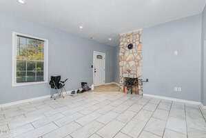 A mostly empty room with light gray walls, white tile floor, a single camping chair, a few personal items near the door, a window, and a corner stone fireplace. A white door and electrical outlets are visible.
