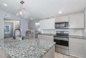 A modern kitchen with gray cabinets, granite countertops, a stainless steel stove and microwave, a double sink with a black faucet, and packaged food items on the counter near the wall.