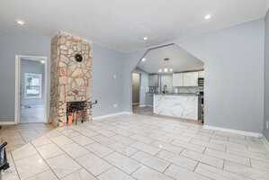 A living area with light-colored tile flooring, light blue walls, a stone fireplace, and a view into a kitchen with white cabinets and a marble-patterned island under modern pendant lights.
