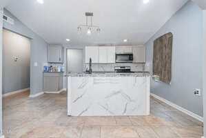 A modern kitchen with light gray walls, a marble-patterned island, stainless steel appliances, white and gray cabinetry, recessed lighting, and large tile flooring. A vent and a wooden accent are visible on the right wall.