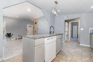 A modern kitchen with a granite island countertop, built-in sink, and dishwasher. The floor is tiled, and there is a laundry area with a stacked washer and dryer visible through an open doorway.