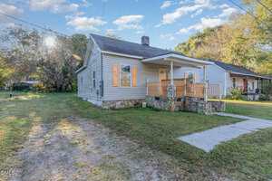 Single-story house with gray siding, orange shutters, and a covered front porch with a wooden railing. The yard is grassy, and a concrete path leads to the porch. Trees and another house are visible in the background.