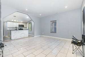 A spacious room with light gray walls and white marble tile flooring, featuring a modern kitchen with white cabinets and a marble island in the background. A black folding chair is placed near a window on the right.