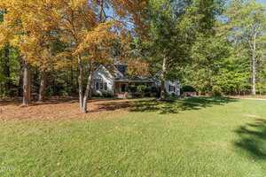 A white house with a front porch is surrounded by trees, some with yellow autumn leaves, and a large grassy lawn under a sunny sky.