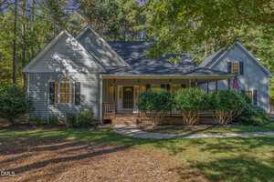 A light gray, single-story house at 120 Marlowe Drive, Youngsville NC 27596 (MLS #10128675), with a covered front porch, white trim, black shutters, mature trees and bushes. An American flag hangs near the entrance and a curved walkway leads to the door.