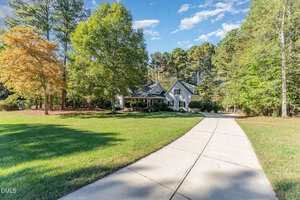 A concrete driveway curves toward a gray house with a front porch, surrounded by green grass and trees under a blue sky with scattered clouds.