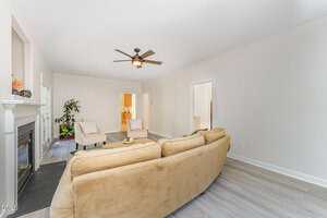 A living room with light-colored walls, a beige sectional sofa, two cream chairs, a fireplace on the left, a ceiling fan, a potted plant, and light wood flooring. Doorways lead to other rooms in the background.