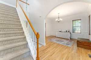 A carpeted staircase with a wooden handrail is on the left, leading up to a second floor. On the right, there is a room with hardwood floors, a rug, a table with a laptop, a chandelier, and windows with blinds.