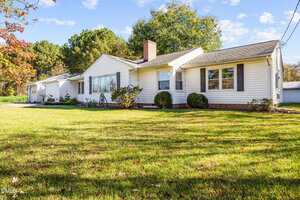 Single-story white house with a gray roof, brick chimney, and attached garage, surrounded by green lawn, shrubs, and trees under a blue sky with scattered clouds.