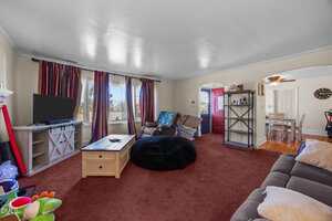A living room with red carpet, beige walls, a TV on a stand, a coffee table, a large black bean bag, a couch, and armchairs. There are large windows with purple curtains and an open view to a dining area.