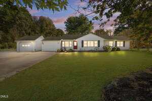 Charming single-story white house at 1155 Bobbitt Road, Kittrell NC 27544, with a red front door, attached garage, large front lawn, and trees in the background. Sunset photo with exterior lights on and a clear sky.