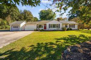 Single-story white house with a red front door, attached garage, large front lawn, and trees in the background under a clear sky. The driveway leads to the garage on the left side of the house.