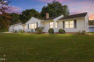 Single-story white house with dark roof, attached garage, shutters, and brick foundation, set on a large green lawn with shrubs in front and trees in the background at sunset.