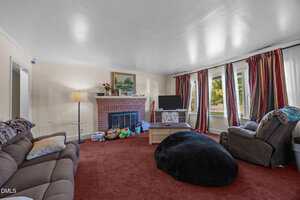 A living room with red carpet, a brown couch, a recliner, a black bean bag, a fireplace with toys and decor on the mantel, a TV on a stand, and large windows with red and white curtains.