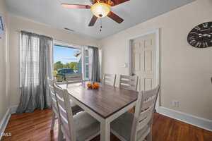 A dining room with a wooden table and six chairs, a ceiling fan with a light, large window with sheer curtains, a wall clock, and hardwood floors. A parked car and greenery are visible outside the window.