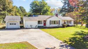 Single-story white house with a red front door, attached garage on the left, and a large concrete driveway. There is a grassy lawn, some small shrubs, and trees in the background under a blue sky.