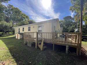 A single-story yellow house with white trim has a wooden deck and stairs leading to a grassy yard. The sky is mostly clear with some clouds and sunlight shining above. Trees surround the house in the background.