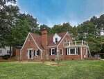 A red brick house at 508 W Lake Road with a steep roof, chimney, and a second-floor balcony, surrounded by grass and trees. The home features black shutters and white trim on the windows and doors.
