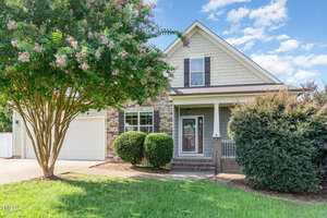 A single-family house at 1731 Riverstone Drive in Franklinton features stone and siding exterior, a covered front porch, attached garage, front shrubs, and a large tree on the left, all set under a blue sky with scattered clouds.
