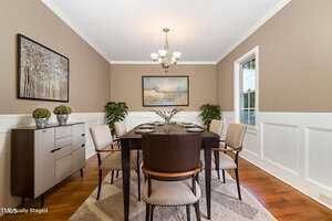 A dining room with a dark wooden table set for six, beige upholstered chairs, a sideboard with decorative plants, two wall paintings, hardwood floor, and a chandelier overhead. White wainscoting lines the tan walls.